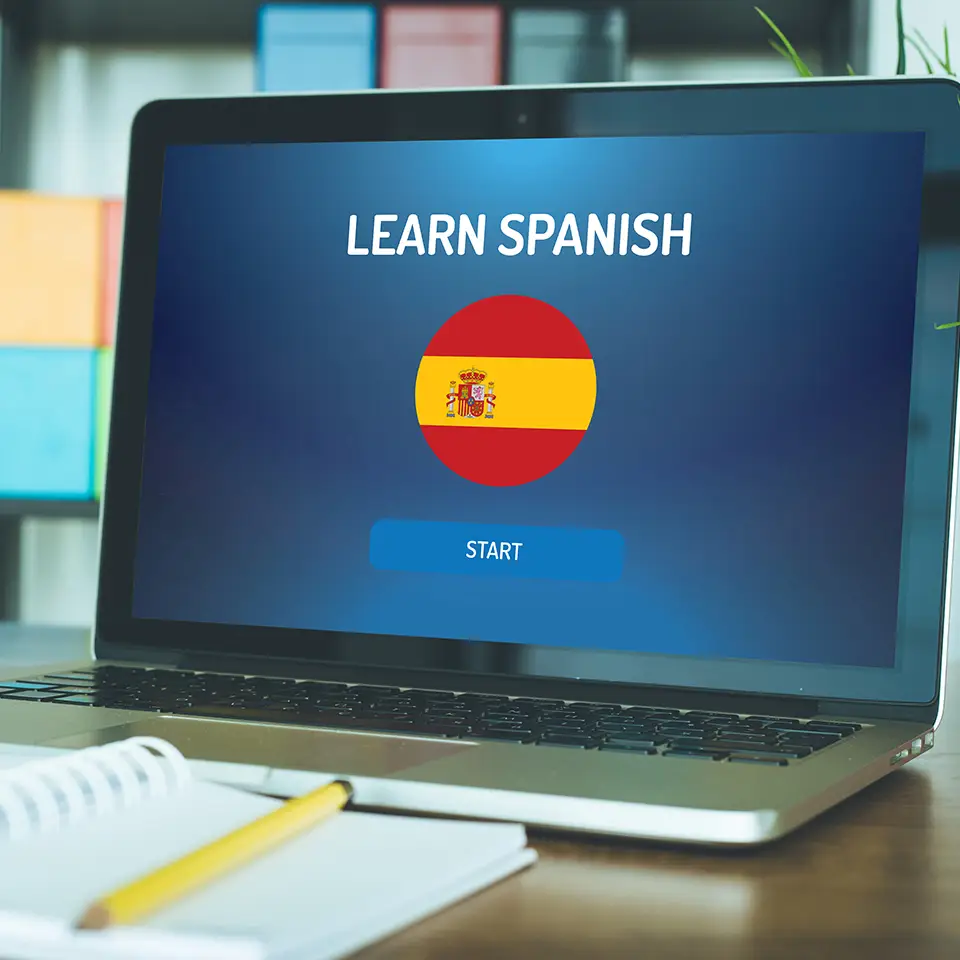 Open laptop displaying a Spanish flag and the words 'Learn Spanish' on the screen. The laptop is one a desk with an open notebook in front of it.